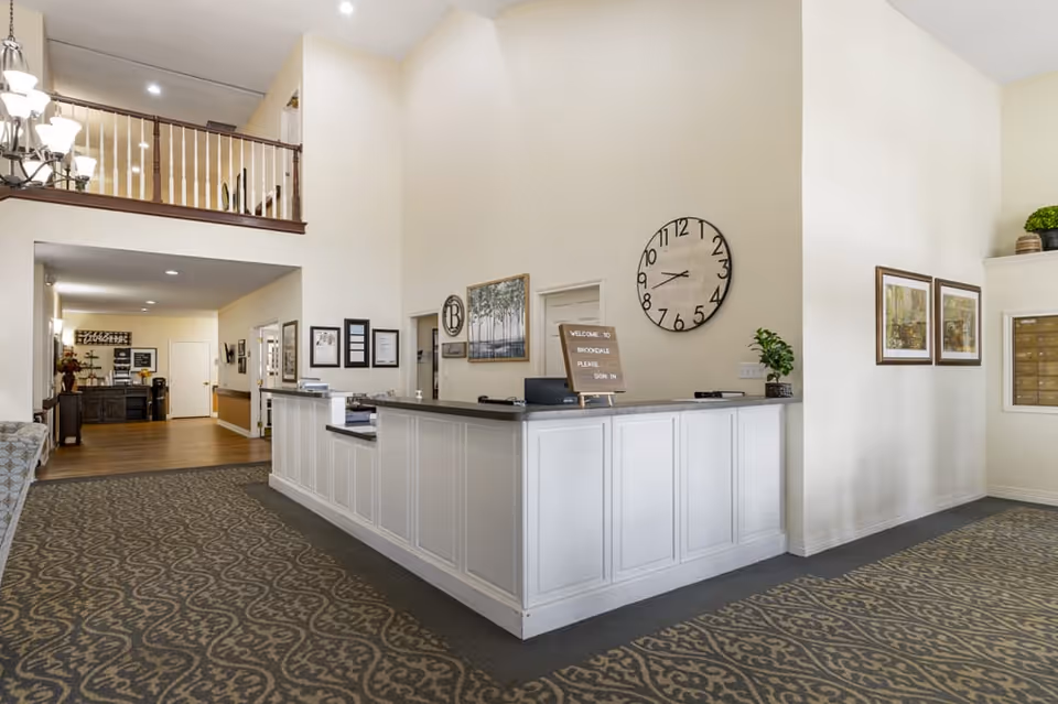 Reception area of Brookdale Bossier City with a white front desk, a large wall clock, framed artwork, and a sign that says 'Welcome to Brookdale Please Sign In'. The space has patterned carpet, light-colored walls, and a high ceiling with a balcony railing above.