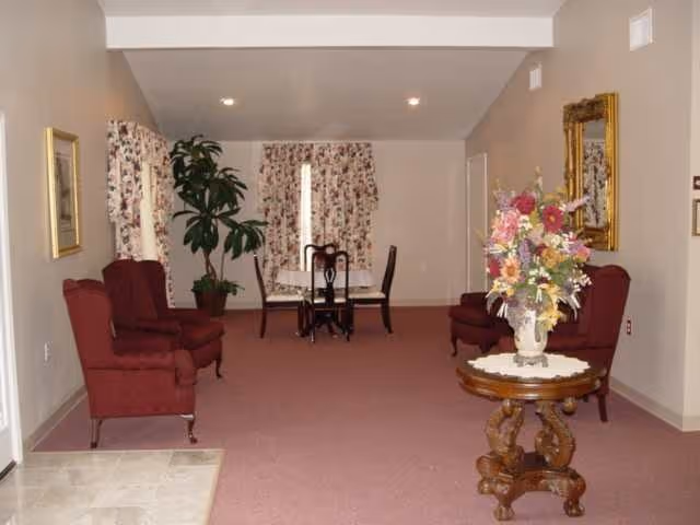 A spacious living room area with maroon upholstered armchairs arranged along the walls, a round wooden table with a floral arrangement in the center, floral curtains on the windows, and a small dining table with chairs in the background.