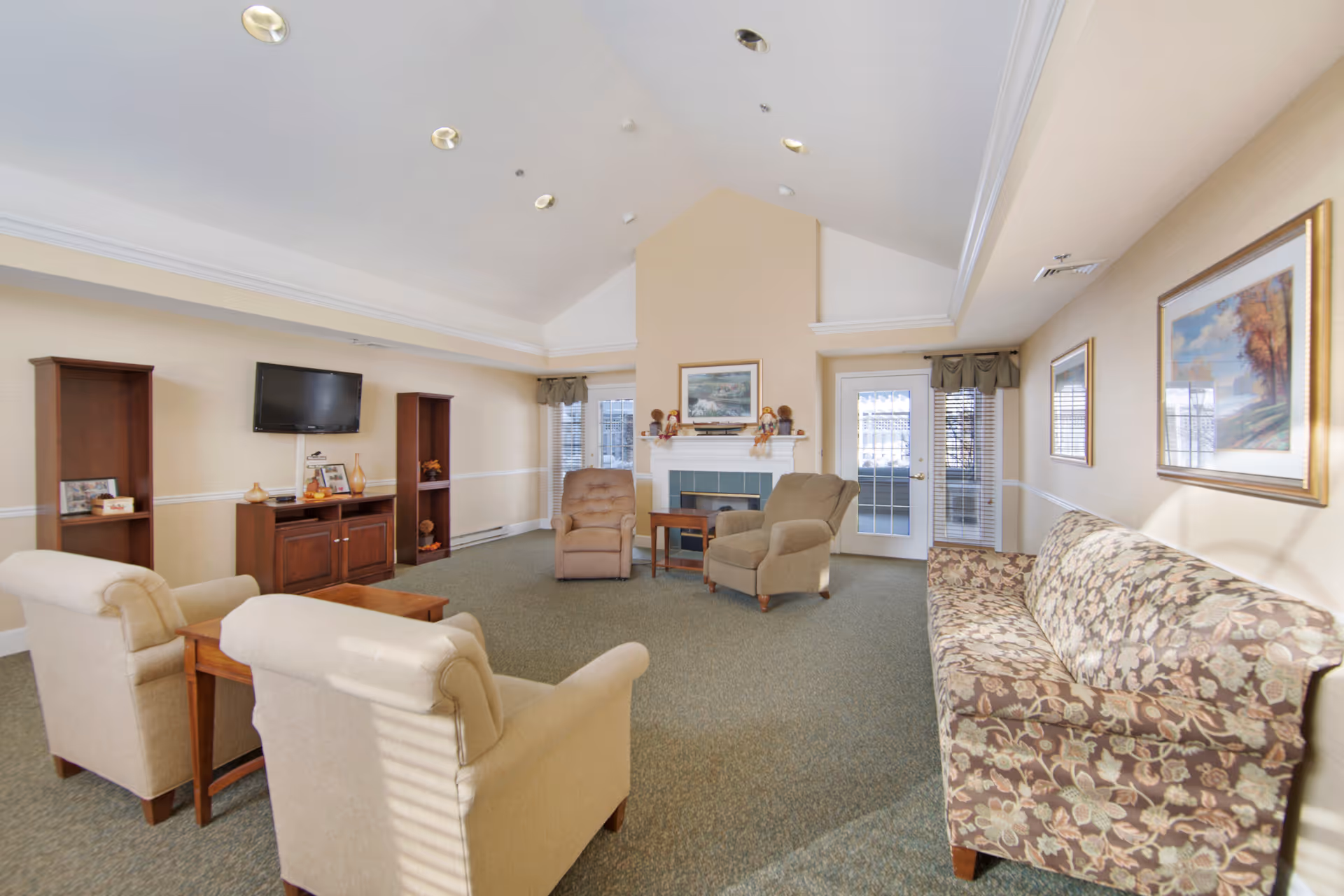 A bright and spacious living room with vaulted ceilings, featuring a floral patterned sofa, two beige armchairs, two brown armchairs, a wooden coffee table, a wooden TV stand with a flat-screen TV mounted above it, and two wooden shelving units. There is a fireplace with decorative items on the mantel and framed artwork above it. The room has windows with blinds and a glass door leading outside.