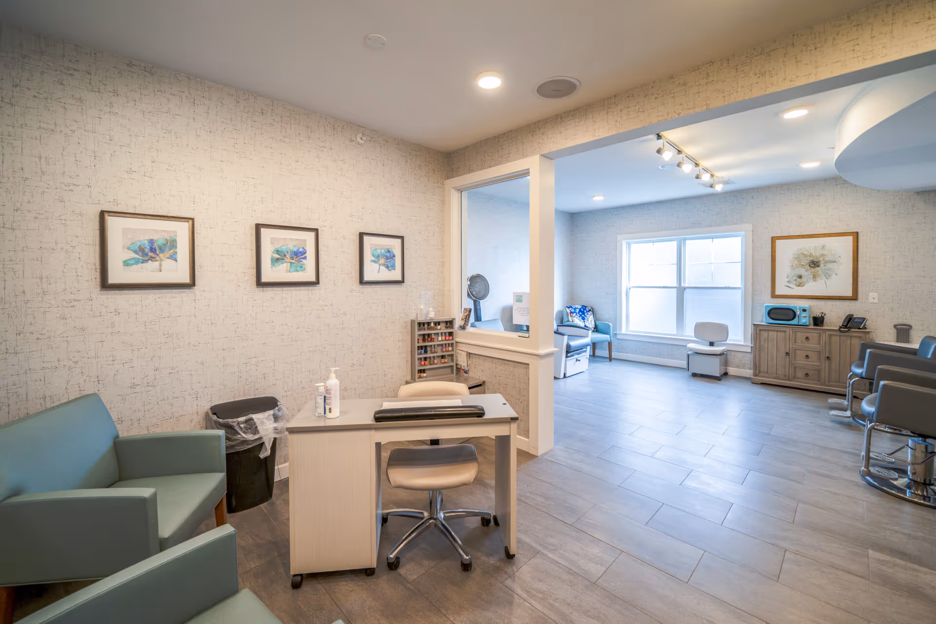 A bright, clean interior room with light-colored walls and tiled flooring. The room features a small desk with a chair, two green armchairs, and a trash bin. On the wall above the armchairs are three framed pictures of blue and green abstract art. Through a large open window-like partition, a larger room is visible with a window letting in natural light, a wooden cabinet with a microwave and phone on top, a framed floral picture on the wall, and several chairs arranged around the space.