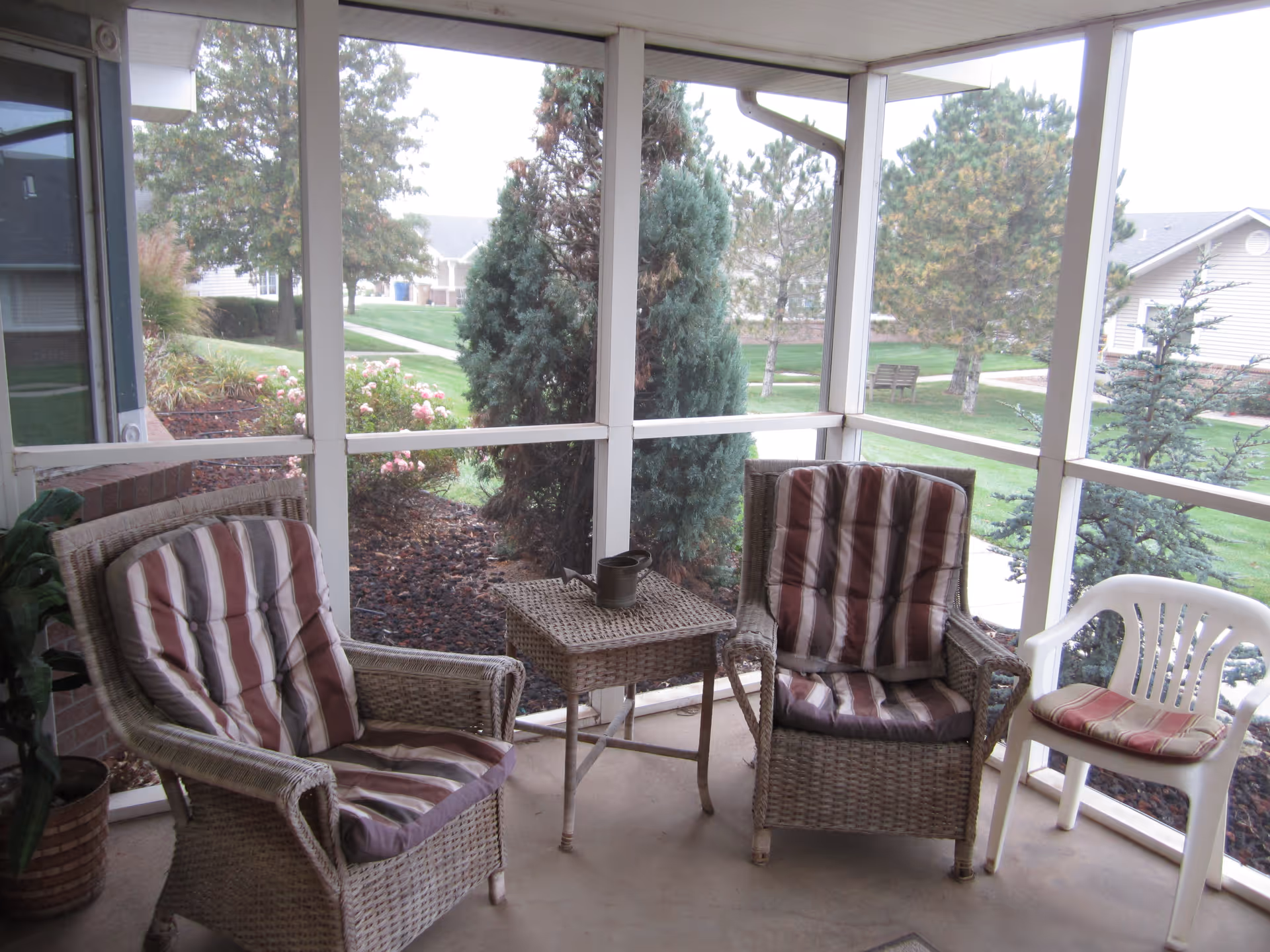 A screened-in porch area with two cushioned wicker chairs and one white plastic chair with a cushion. A small wicker table with a watering can sits between the two wicker chairs. Outside the porch, there are trees, bushes, and a grassy area with a sidewalk and residential buildings in the background.