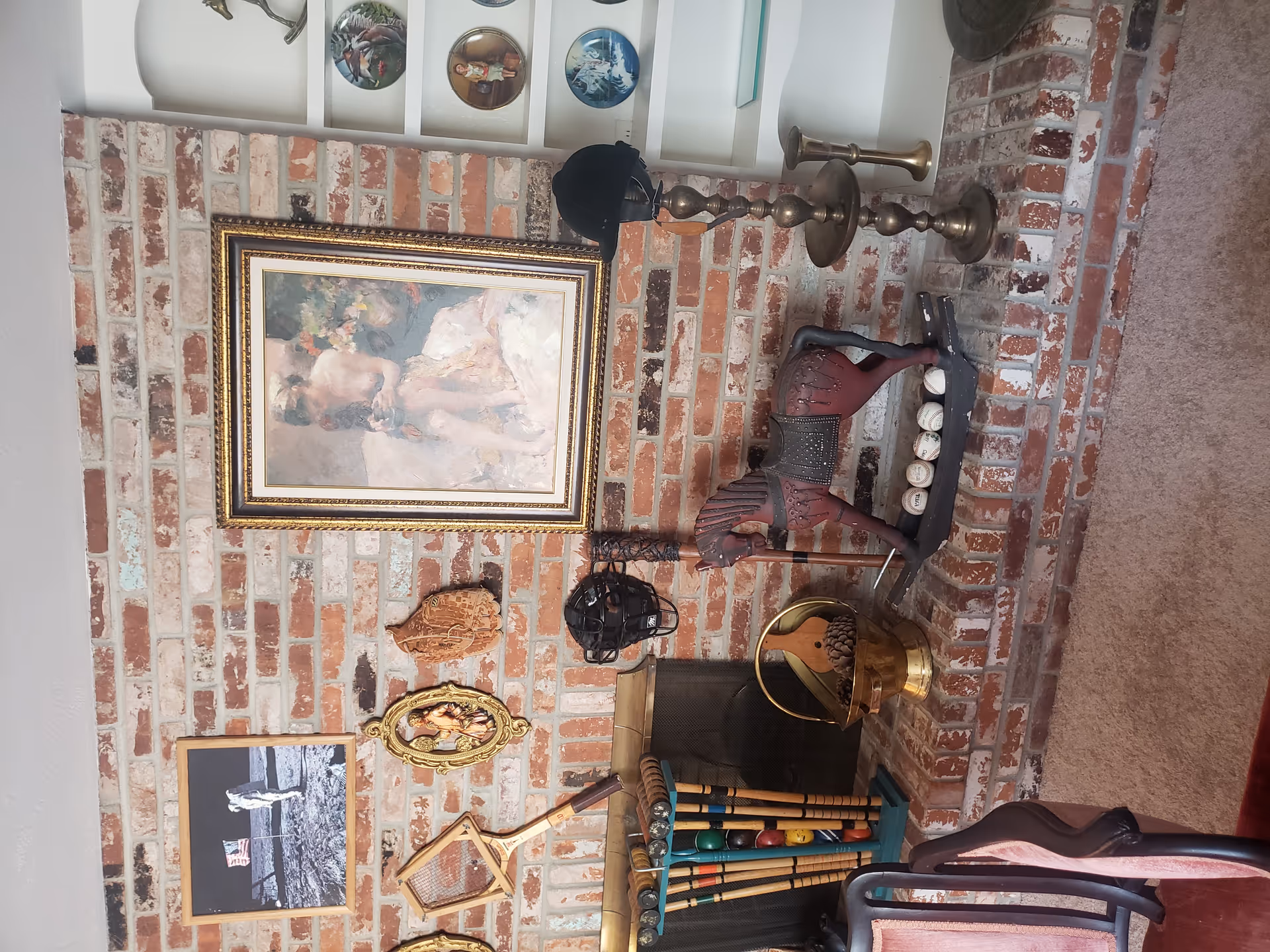 A cozy living room corner with a brick fireplace decorated with various vintage items including a wooden rocking horse, baseballs, a baseball glove, a catcher's mask, croquet mallets, and framed artwork on the brick wall. To the right, there is a white built-in shelf displaying decorative plates and a brass candlestick holder with a black hat on top.