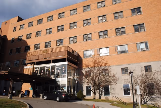 Exterior view of a multi-story brick building with numerous windows, a covered entrance with glass walls decorated with flower stickers, a car parked in front, and leafless trees near the entrance under a partly cloudy sky.