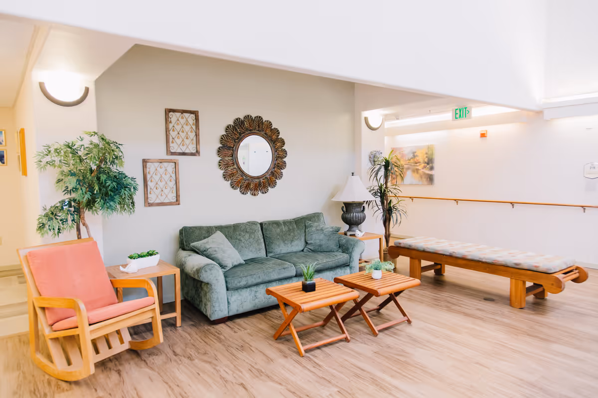 A bright and spacious living room area in Woodland Heights Assisted Living featuring a green upholstered sofa with cushions, a wooden rocking chair with a pink cushion, two small wooden coffee tables with potted plants, a long cushioned bench, decorative wall art including a round mirror and framed pieces, a table lamp on a side table, and indoor plants. The room has light wood flooring and soft lighting.
