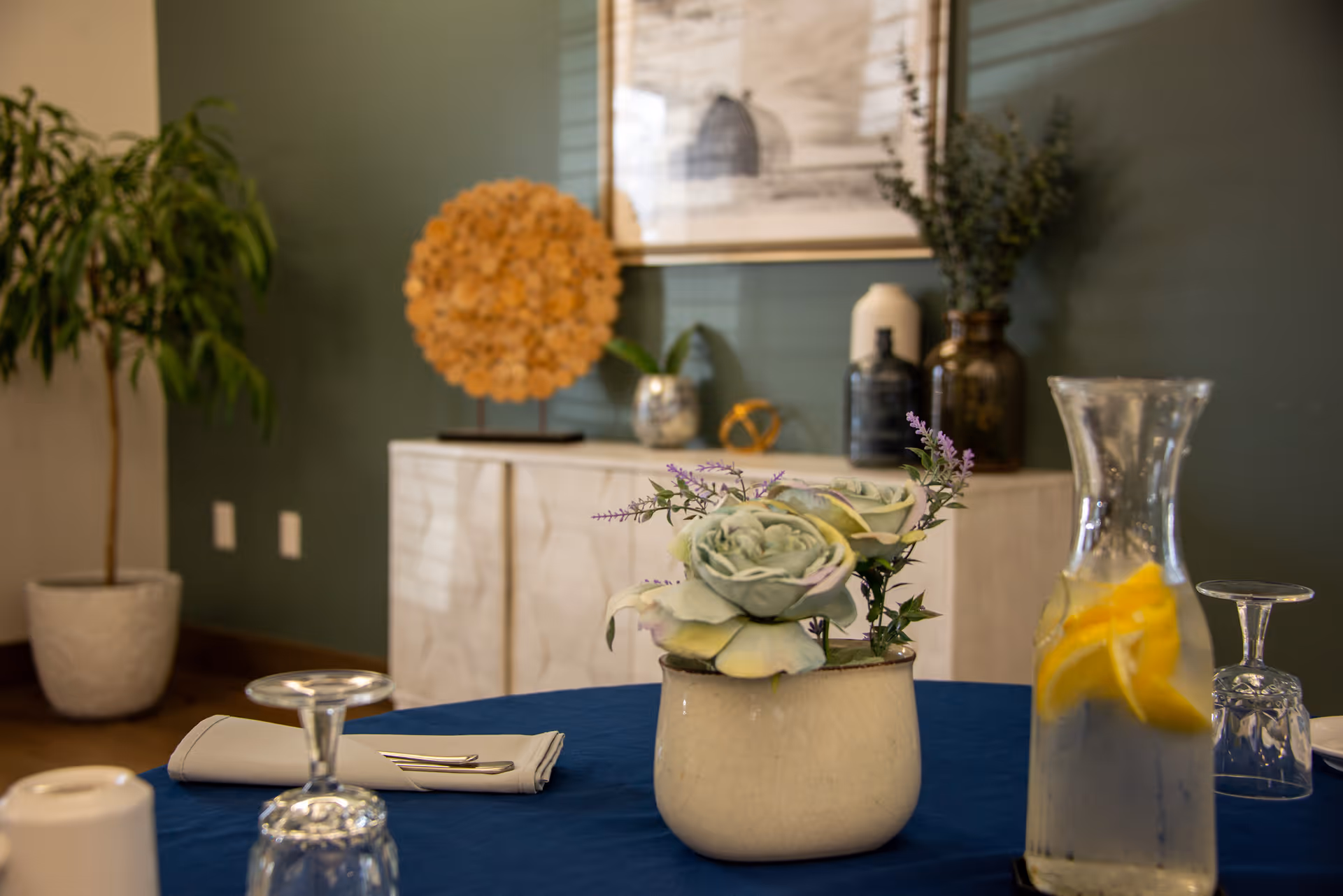 Table set with a floral centerpiece, water pitcher with lemon slices, glasses and napkins in a decorated dining area.