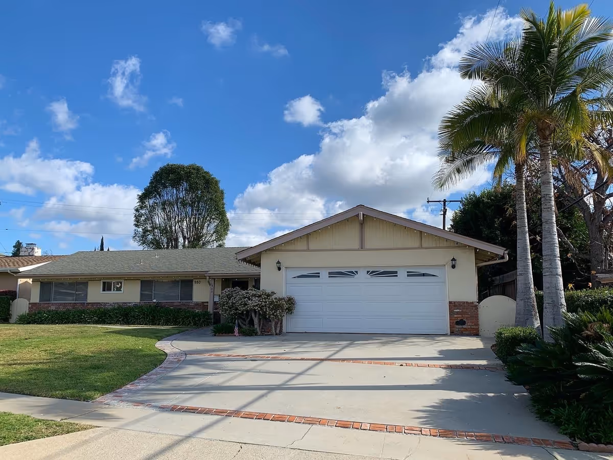 Single-story residential building with a two-car garage, a well-maintained front lawn, palm trees, and a clear blue sky with scattered clouds.