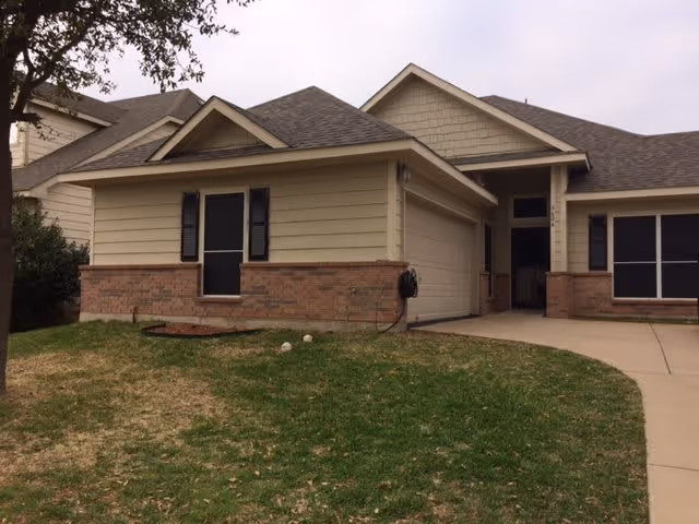Single-story residential building with beige siding and brick accents, a garage, and a driveway. There is a small grassy lawn in front with a tree on the left side under an overcast sky.