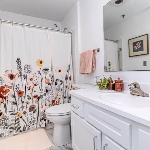 A clean bathroom featuring a white toilet, a white vanity with a sink, a large mirror above the sink, and a shower with a floral-patterned curtain. A pink towel hangs on a towel rack next to the mirror, and there are decorative items including a small plant and amber-colored containers on the vanity countertop. A framed floral picture is visible on the wall reflected in the mirror.