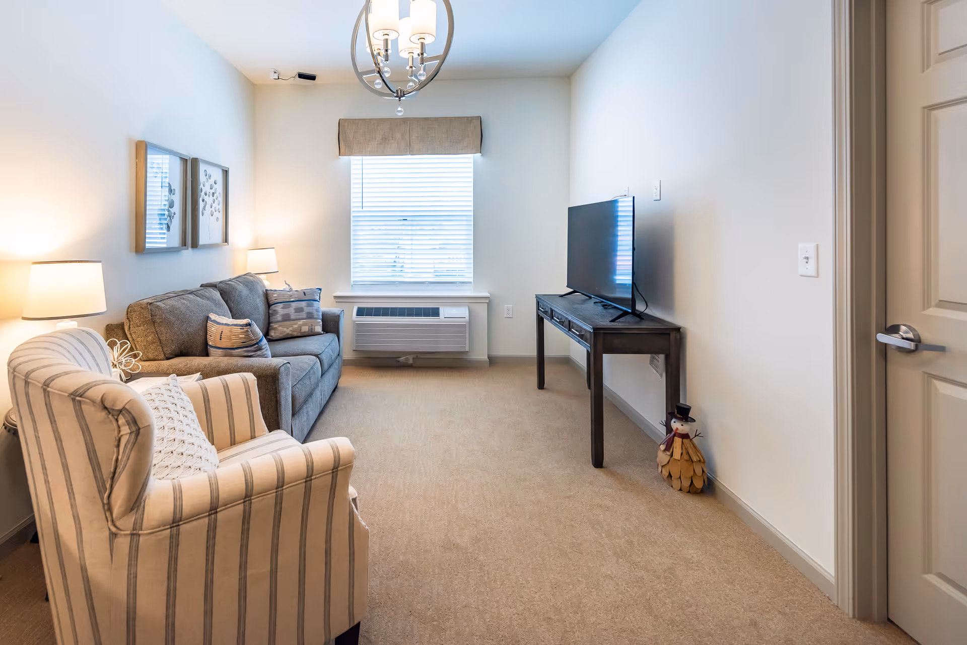 A cozy living room with a striped armchair and a gray sofa adorned with cushions. There are two table lamps on either side of the sofa, a window with blinds and a valance, a flat-screen TV on a dark wooden console table, and a small decorative snowman figure on the floor near the door.