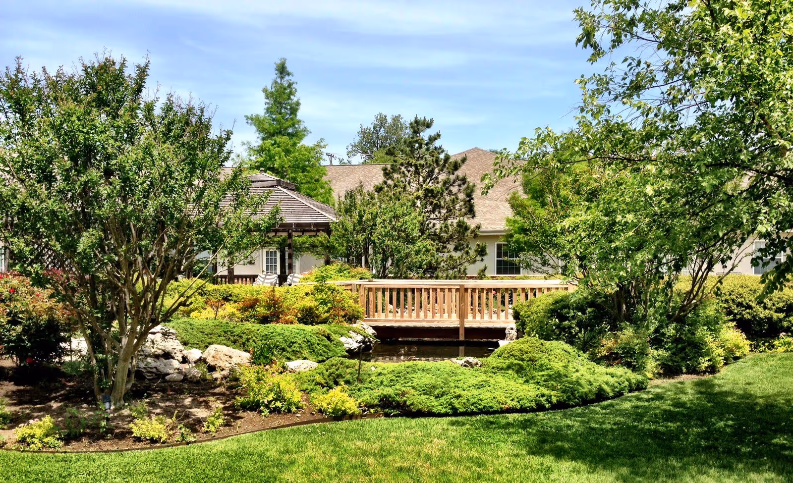 A well-maintained garden area with lush green grass, various shrubs, and trees. There is a wooden bridge over a small pond or water feature, and a gazebo structure is visible in the background. The sky is clear with some clouds, and a building with a beige roof is partially visible behind the greenery.