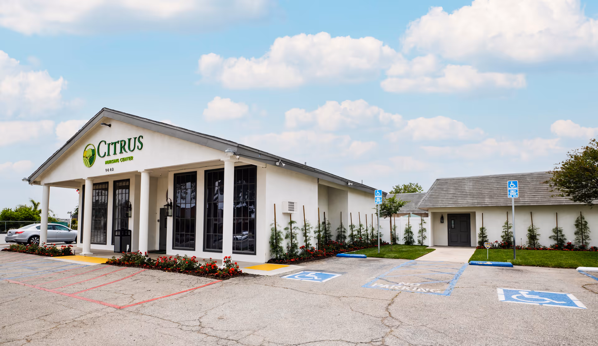 Exterior view of Citrus Nursing Center building with white walls, large windows, and a covered entrance supported by columns. There are several handicap parking spaces in front, a few cars parked, and landscaping with bushes and flowers around the building. The sky is partly cloudy.