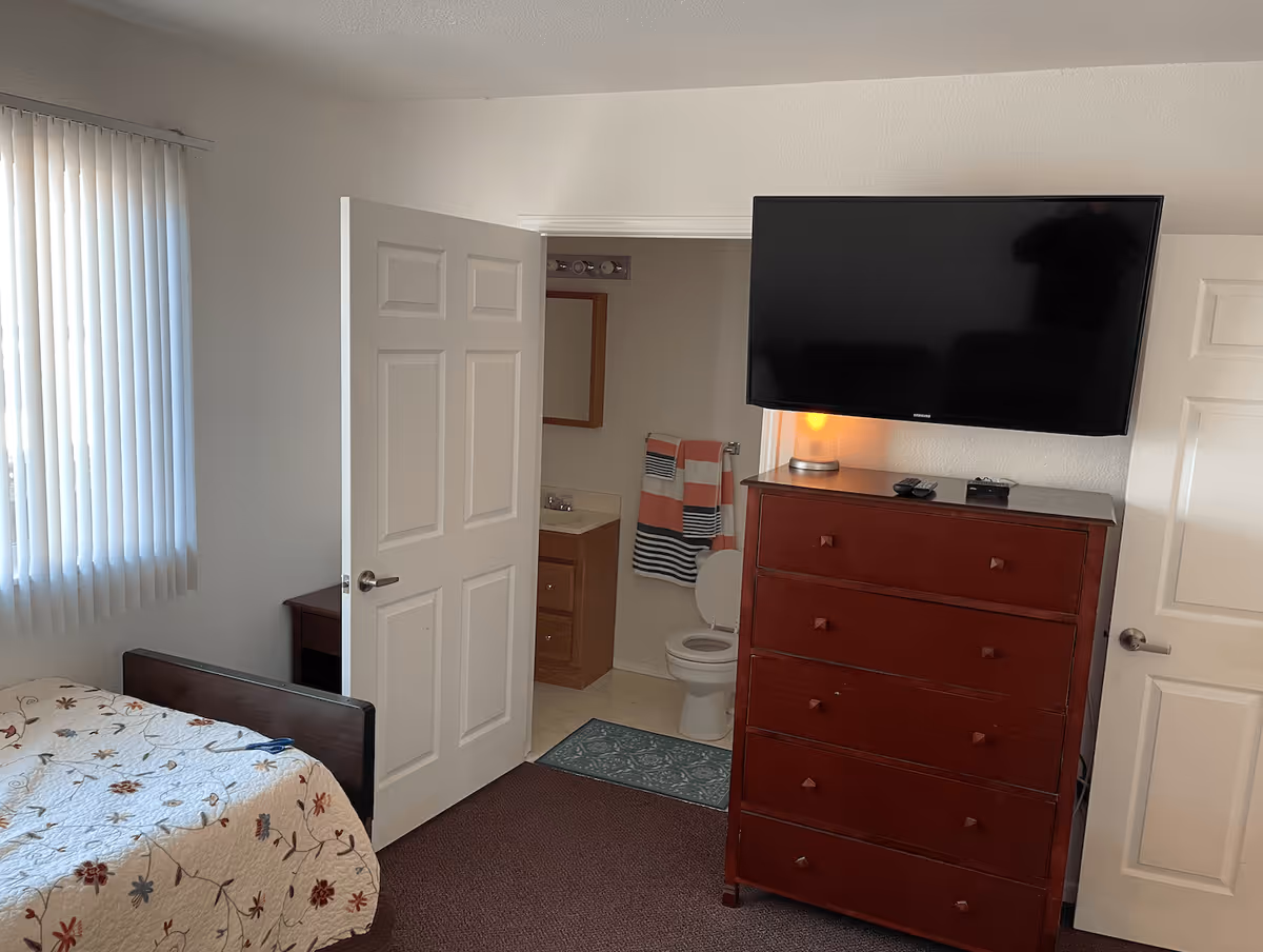 A bedroom with a bed covered in a floral quilt, a window with vertical blinds, a wooden dresser with a flat-screen TV mounted above it, and an open door leading to a bathroom with a toilet, sink, and striped towels.