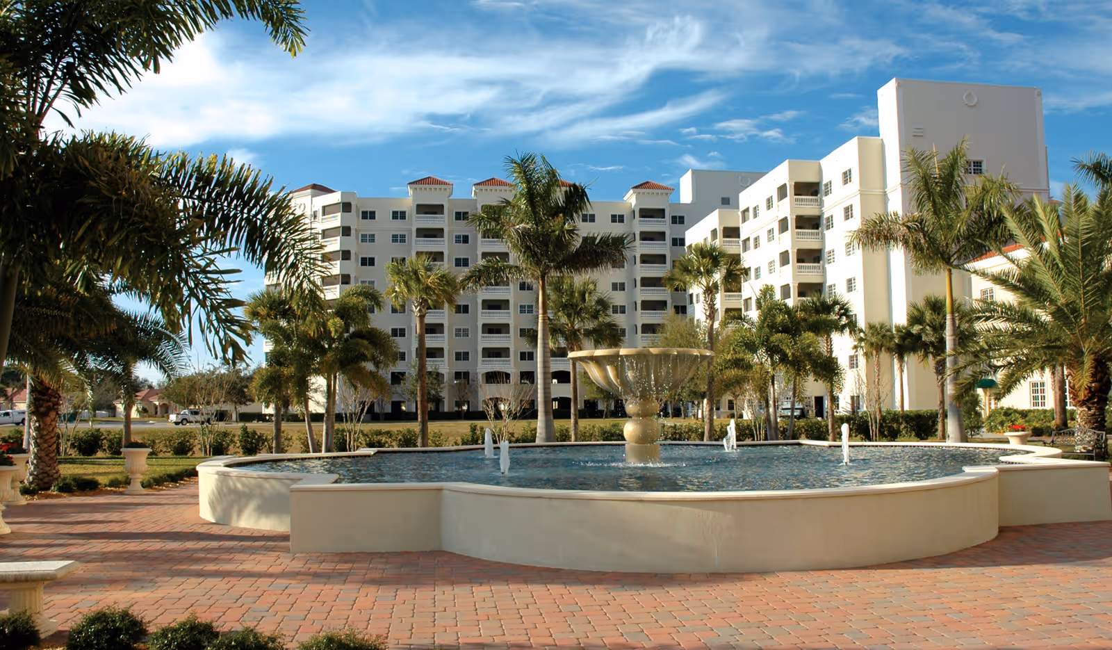 Outdoor view of a senior living facility named Jacaranda Trace featuring a large decorative water fountain surrounded by palm trees and a paved brick walkway, with a multi-story white building in the background under a partly cloudy blue sky.