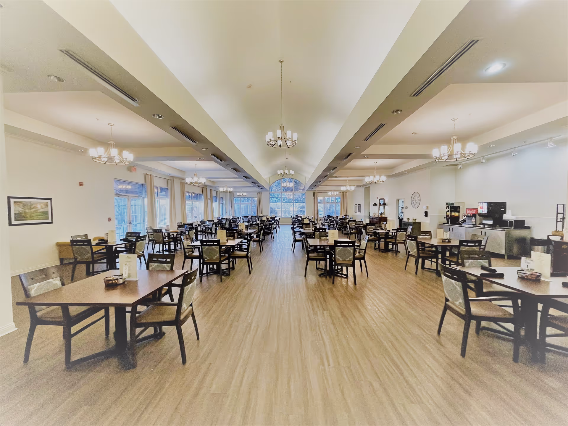 A spacious dining room with multiple wooden tables and chairs arranged neatly. The room has large windows on the left side letting in natural light, chandeliers hanging from the ceiling, and a beverage station on the right side. The floor is wooden, and the walls are painted white.