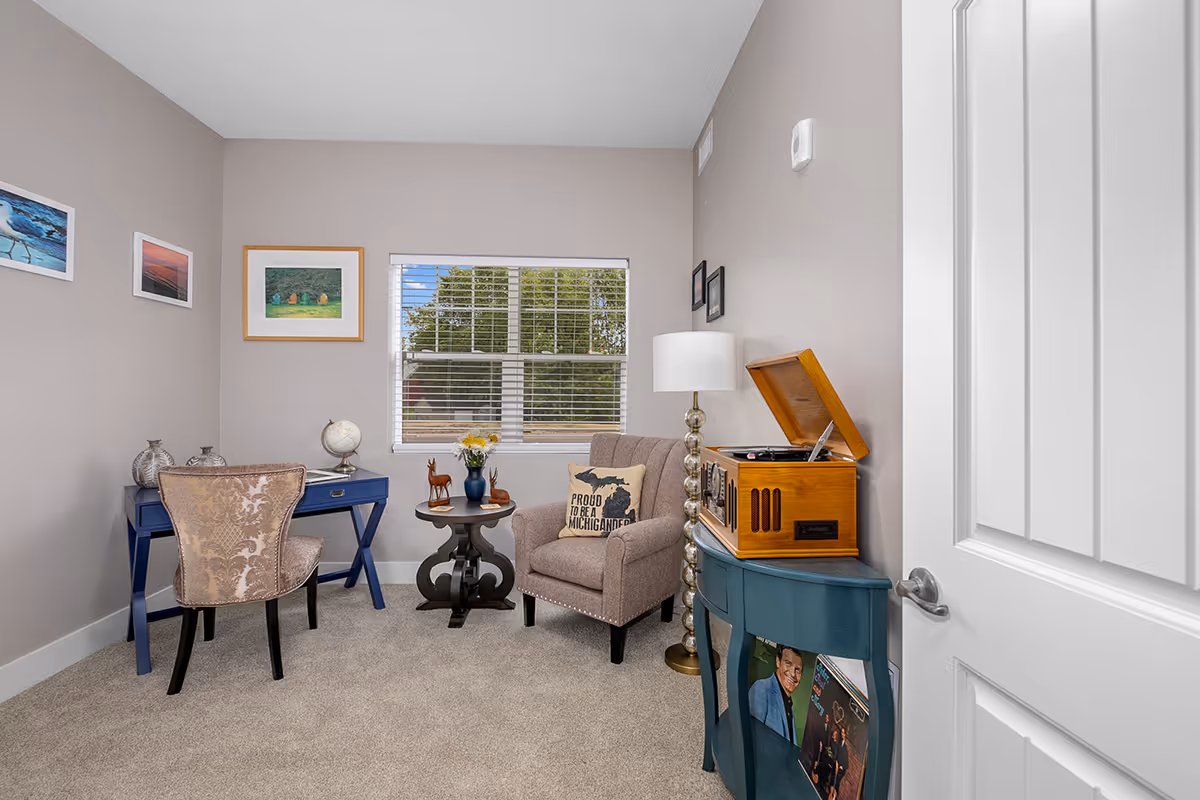 A cozy room with beige walls and carpet featuring a blue desk with a patterned chair, a small round table with decorative items, a comfortable armchair with a pillow that reads 'Proud to be a Michigander,' a tall floor lamp, and a blue cabinet holding a vintage record player and vinyl records. A window with white blinds shows green trees outside, and framed pictures hang on the walls.