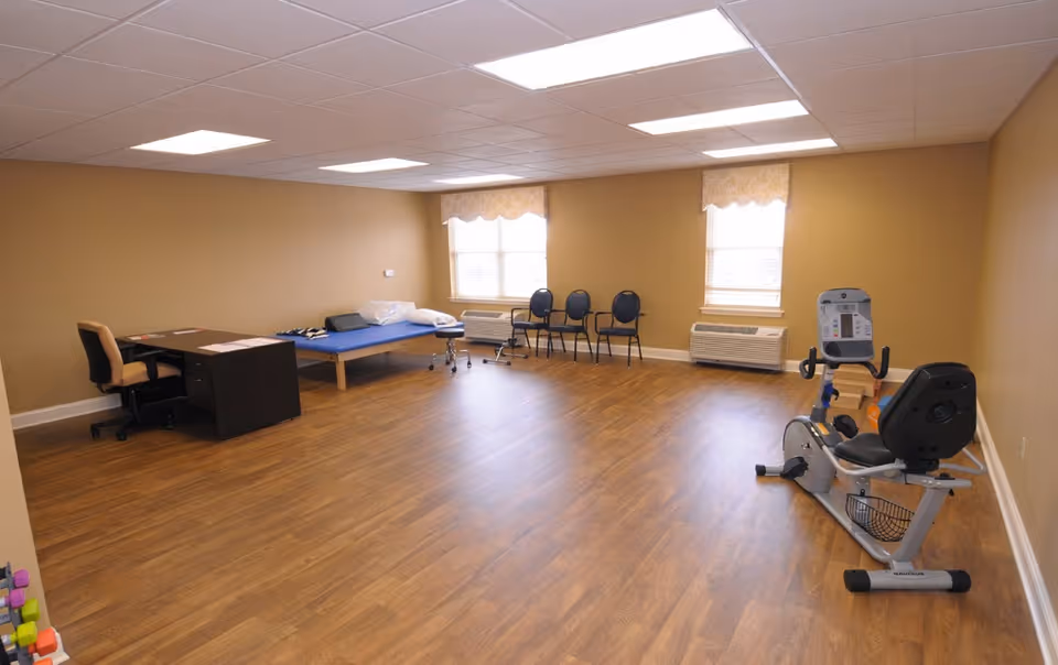 A spacious interior therapy/exercise room with wood floors, a recumbent exercise bike, treatment table, desk, and chairs.
