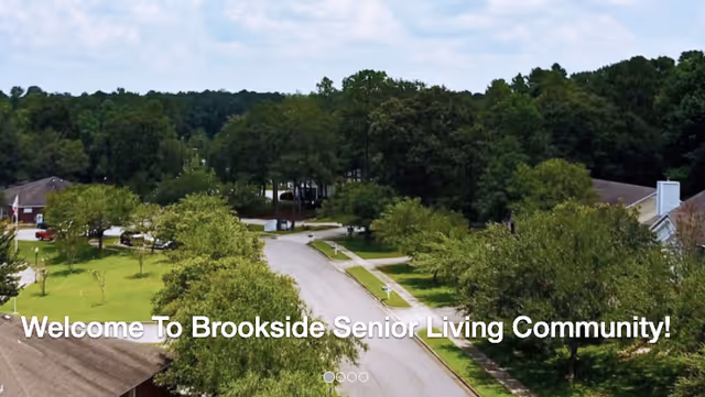 Aerial view of a senior living community street lined with trees and houses, with a welcome message overlay that reads 'Welcome To Brookside Senior Living Community!'