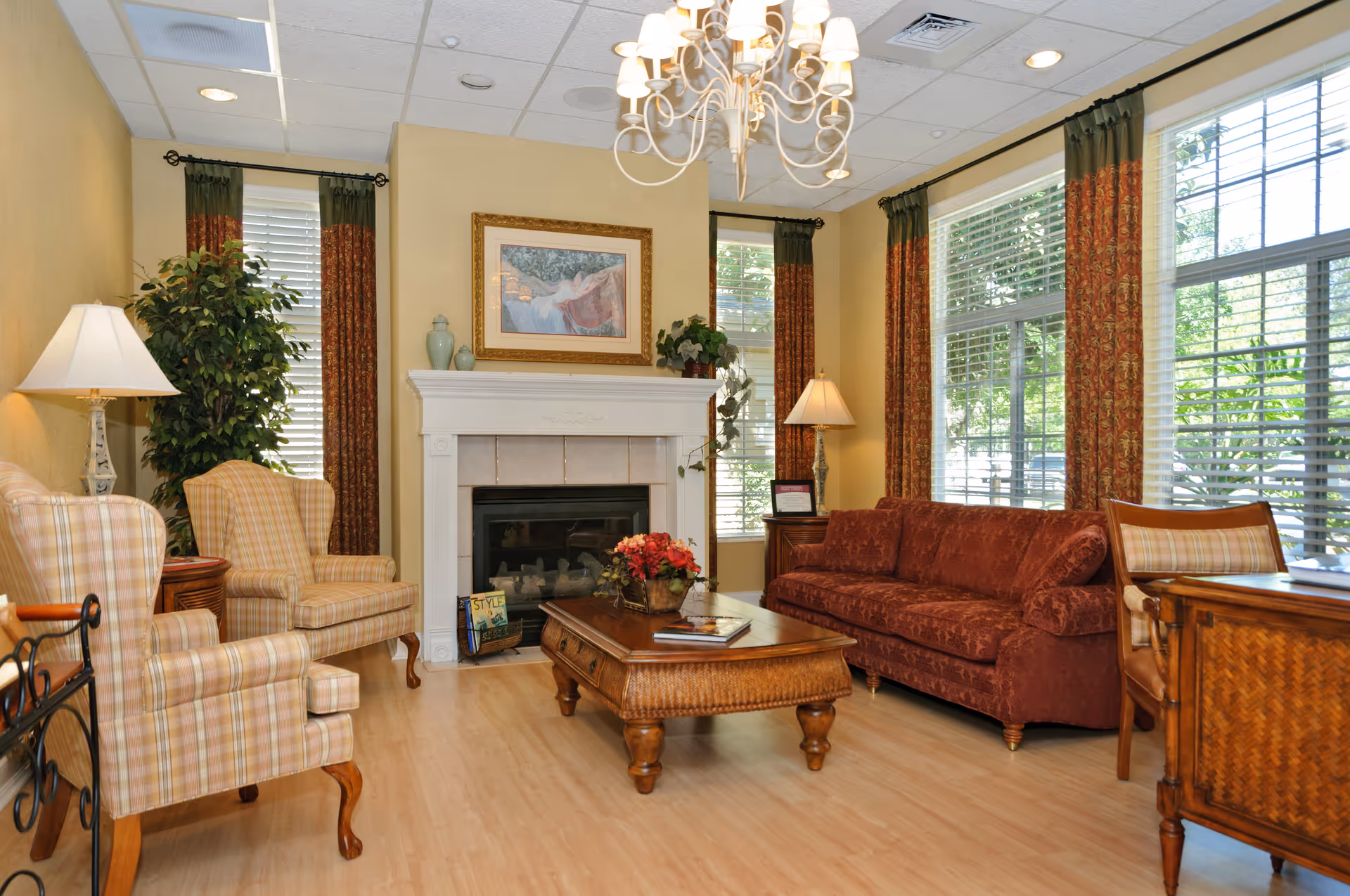 A cozy living room with a red sofa, two plaid armchairs, a wooden coffee table with a flower arrangement, a fireplace with decorative vases and a framed painting above it, large windows with patterned curtains, two table lamps, and a chandelier hanging from the ceiling.