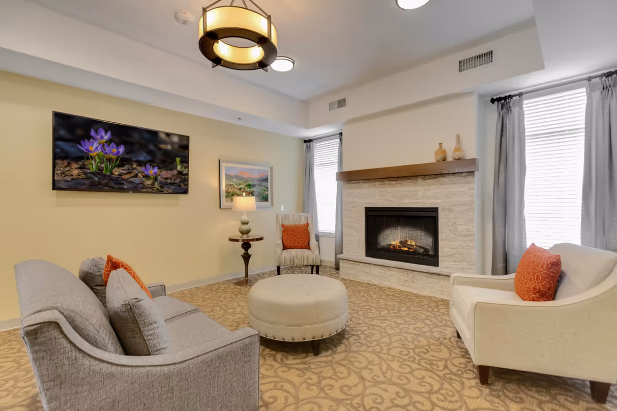 Cozy memory care living room with upholstered chairs, a round ottoman, a wall-mounted TV, and a lit fireplace.