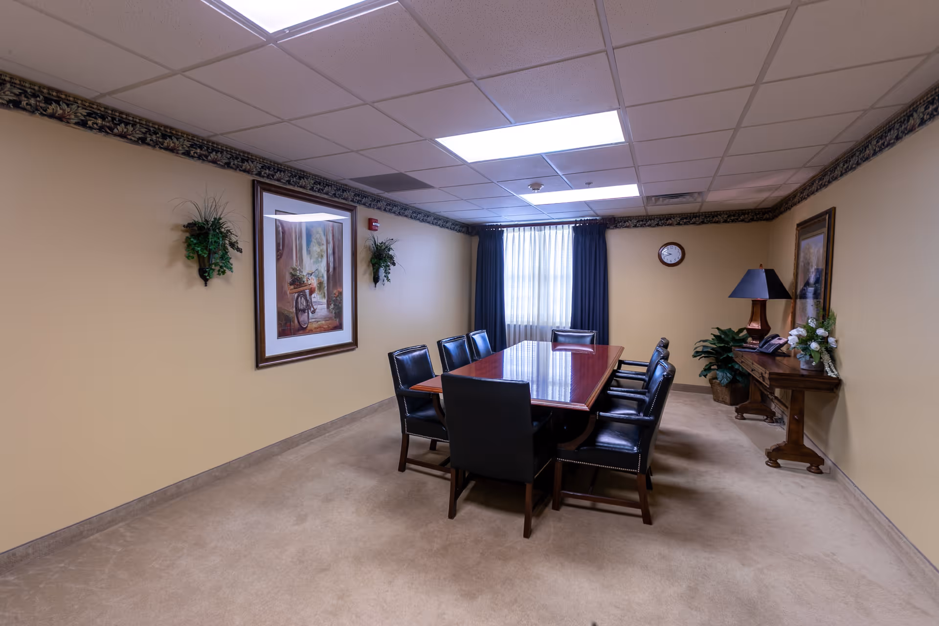 A conference room with a long wooden table surrounded by eight black leather chairs. The room has beige walls with decorative border trim near the ceiling, a window with dark curtains, two framed paintings, two wall-mounted plants, a clock, and a wooden side table with a lamp, a phone, and a flower arrangement.
