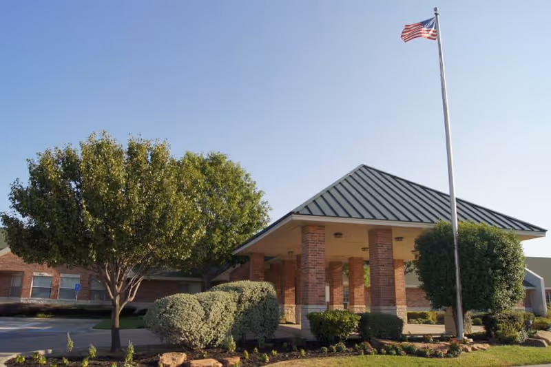 Exterior view of a single-story brick building with a green metal roof, surrounded by neatly trimmed bushes and trees. An American flag flies on a tall flagpole near the entrance under a covered driveway.