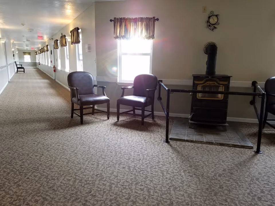 A long carpeted hallway with multiple windows on the left side, each with brown curtains. Two brown armchairs are placed near a window on the right side, next to a black wood-burning stove with a protective railing around it. A clock is mounted on the wall above the stove.