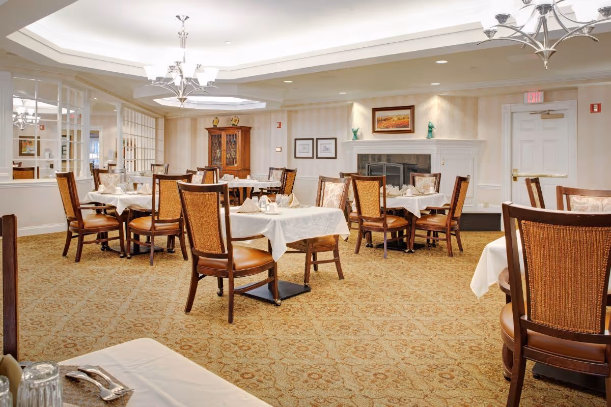 Well-lit dining room with multiple tables set with white linens, wooden chairs, a fireplace, and chandeliers.