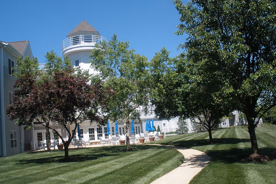 Outdoor view of Cape Albeon Assisted Living facility showing a well-maintained lawn with a curved concrete walkway, several trees providing shade, and a building with a distinctive white tower and blue patio umbrellas on the seating area.