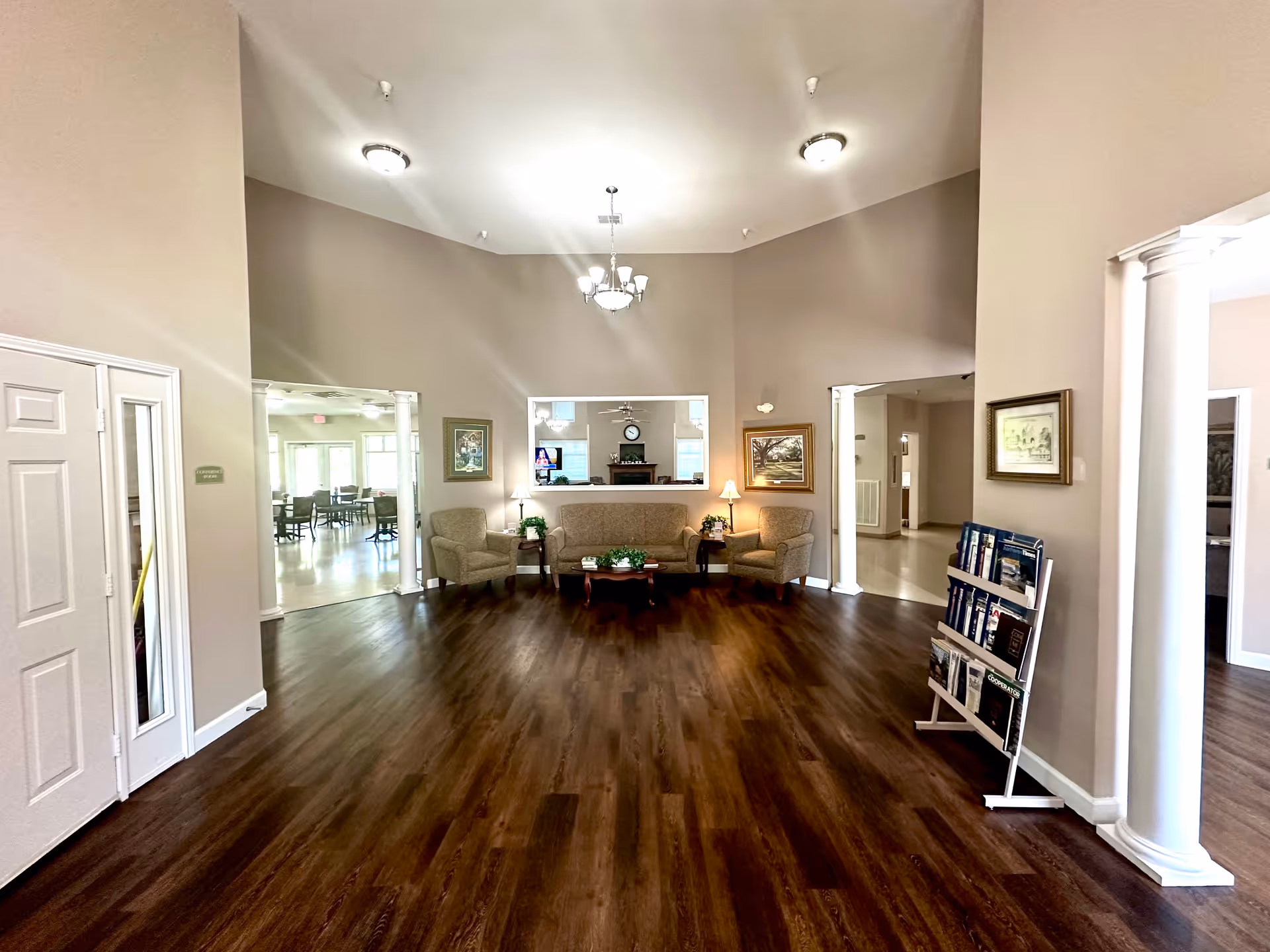 A spacious and well-lit common area in an assisted living facility with wooden flooring, beige walls, and a high ceiling. The room features a beige sofa and two armchairs arranged around a wooden coffee table with plants and books. There are framed paintings on the walls and a chandelier hanging from the ceiling. Two open doorways with white columns lead to adjacent rooms, one of which appears to be a dining area with tables and chairs. A magazine rack is positioned near one of the doorways.