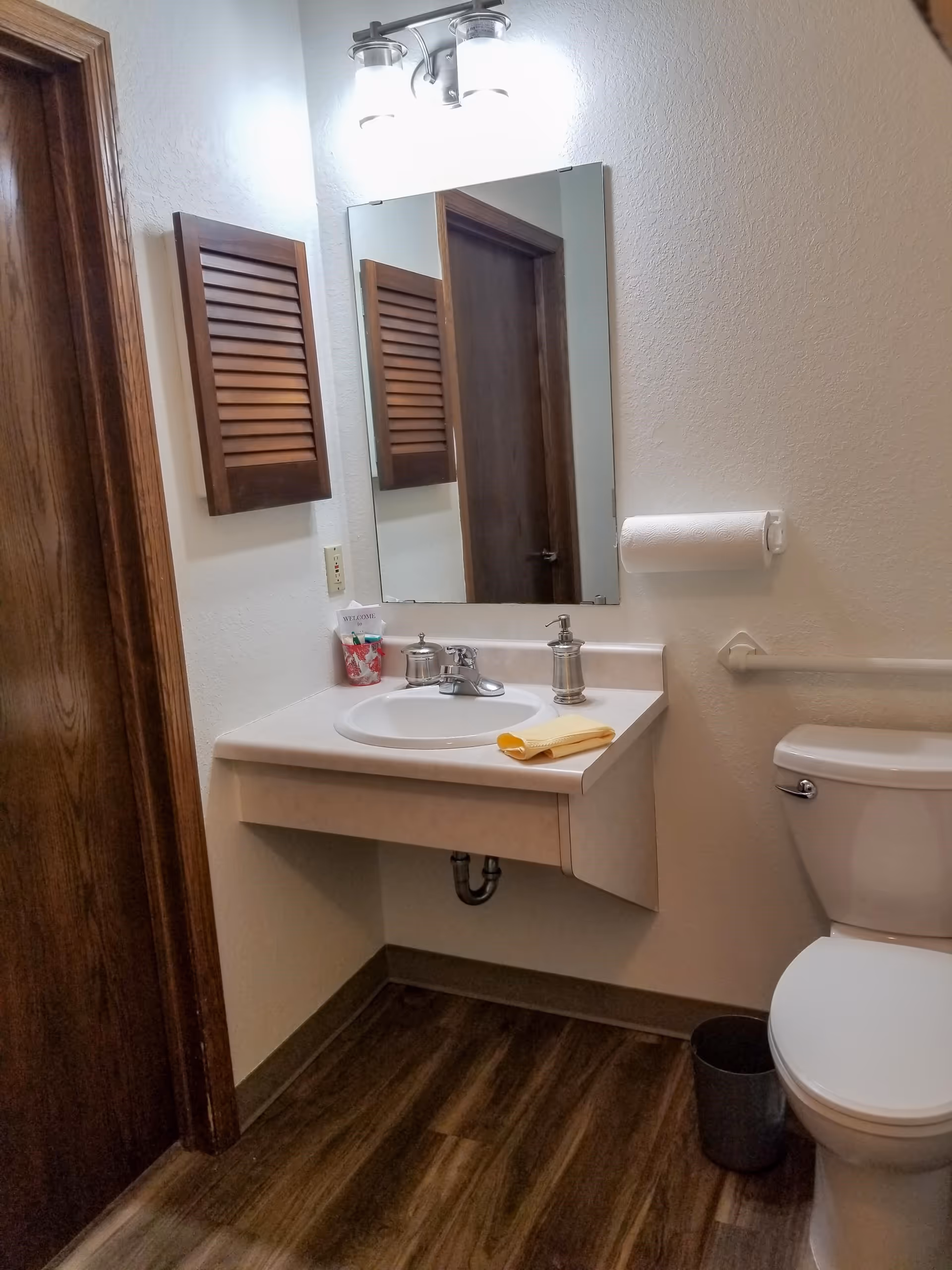 A small bathroom with a white sink and countertop, a mirror above the sink, a wooden cabinet on the wall, a toilet with a grab bar next to it, a paper towel holder with a roll of paper towels, and a small trash can on the floor. The floor has wood-like tiles and the walls are painted white.