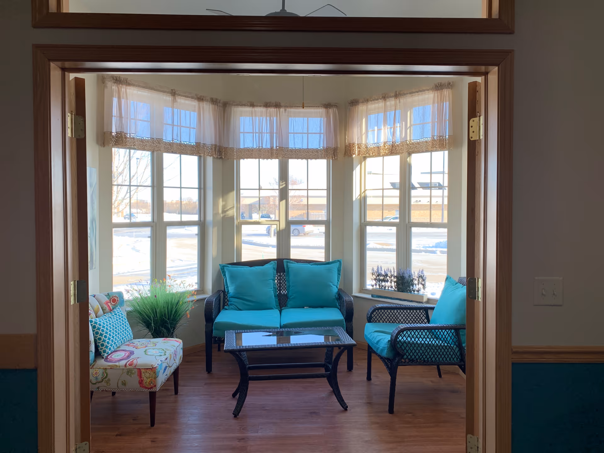 A cozy sitting area with a wicker loveseat and chair with turquoise cushions, a glass-top coffee table, and a floral patterned accent chair. The space is surrounded by large windows with sheer curtains, letting in natural light and showing a snowy outdoor scene.