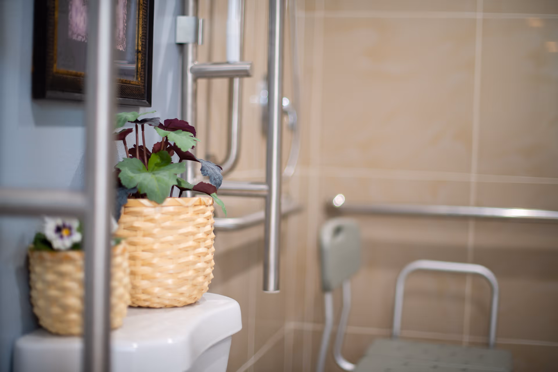 Close-up view of a bathroom corner showing a white toilet tank with two woven baskets containing artificial plants on top. The background features beige tiled walls, a metal grab bar, and a shower chair with a backrest, indicating an accessible bathroom setup.