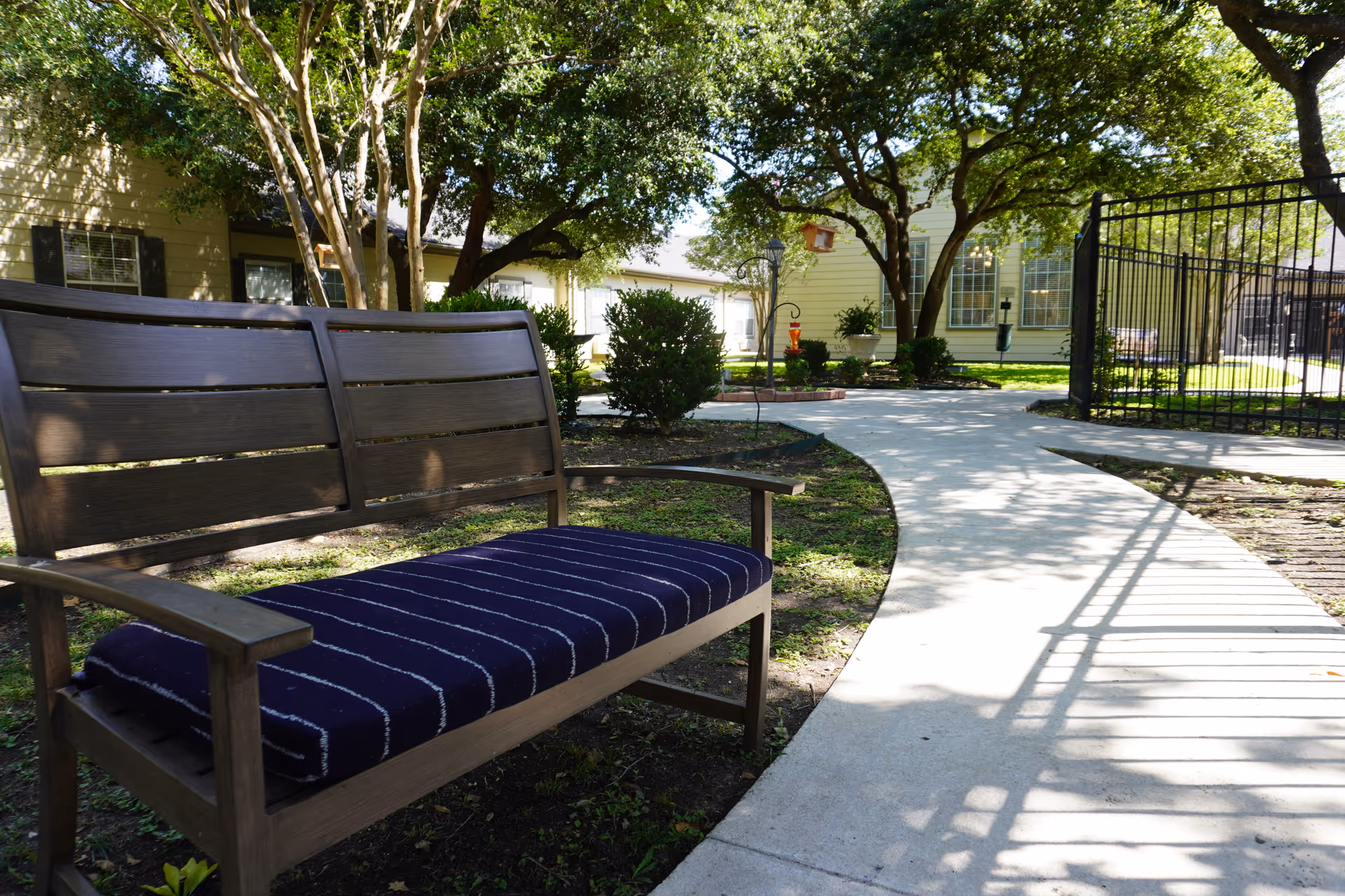 A wooden bench with a dark blue cushion featuring white stripes is placed beside a curved concrete pathway in a garden area. The scene includes green trees, bushes, and a yellow building with large windows in the background. A black metal fence is visible on the right side of the pathway.