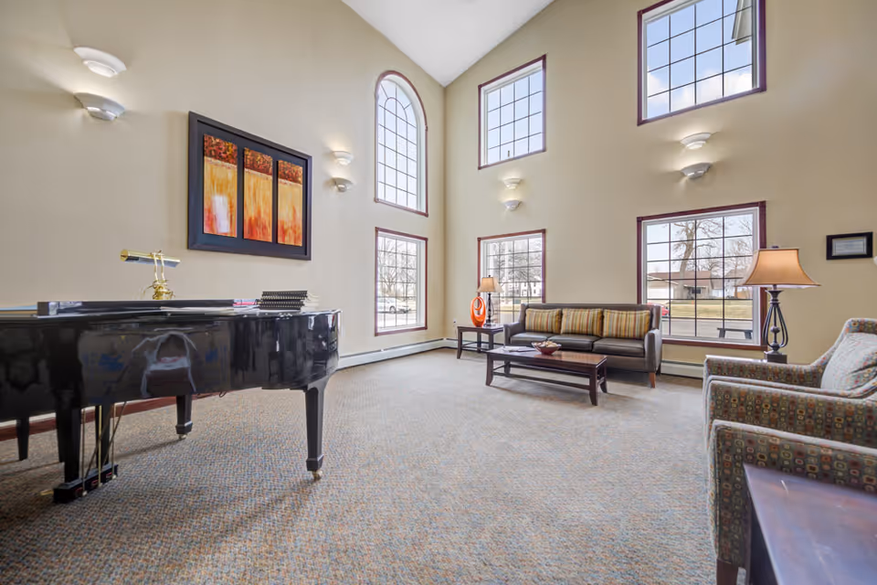 A spacious living room with high ceilings and large windows letting in natural light. The room features a black grand piano on the left, a brown sofa with striped cushions, two patterned armchairs, a coffee table, side tables with lamps, and colorful abstract artwork on the wall.