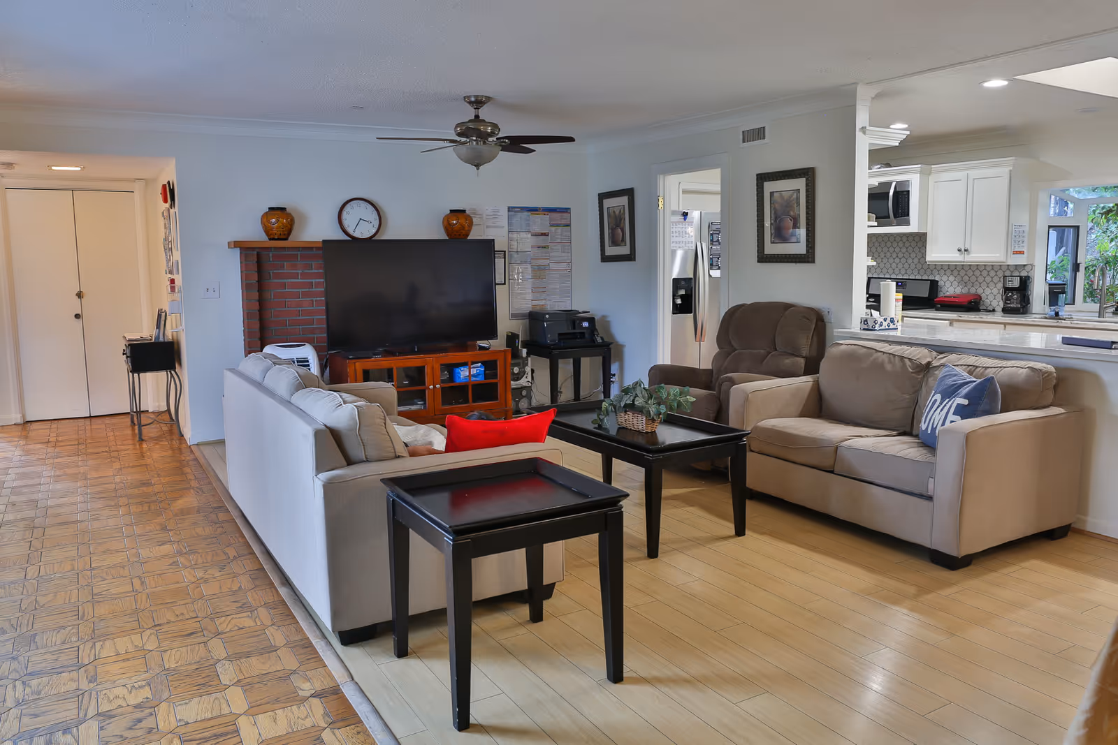 A cozy living room area in a senior care facility featuring a beige sofa with a blue pillow, a brown recliner chair, a black coffee table with a plant, and a large flat-screen TV on a wooden stand against a wall with a clock and decorative vases. The room has light wood flooring, a ceiling fan, and an open view into a white kitchen with modern appliances.