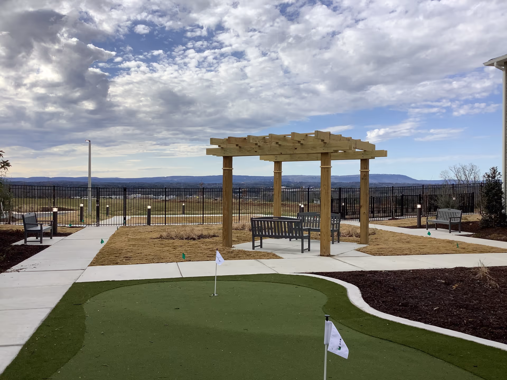 Outdoor courtyard with a wooden pergola, benches and a small putting green overlooking a fenced landscape and distant hills under a cloudy sky.