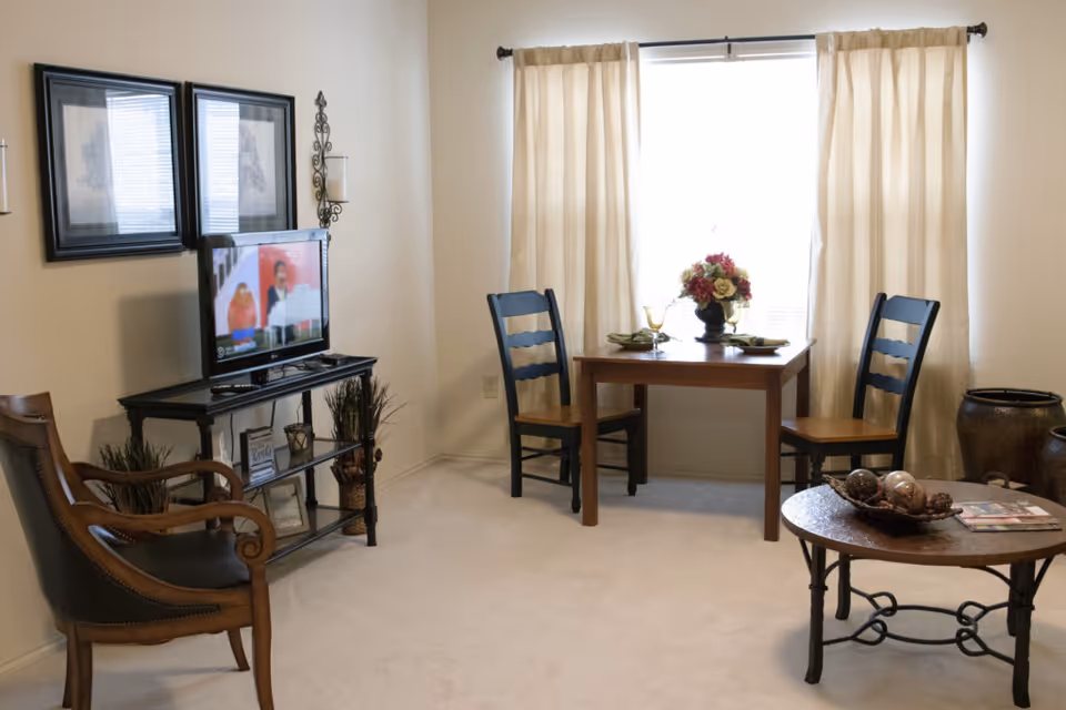 Well-lit living room with a TV on a stand, two chairs and a small dining table by a curtained window.