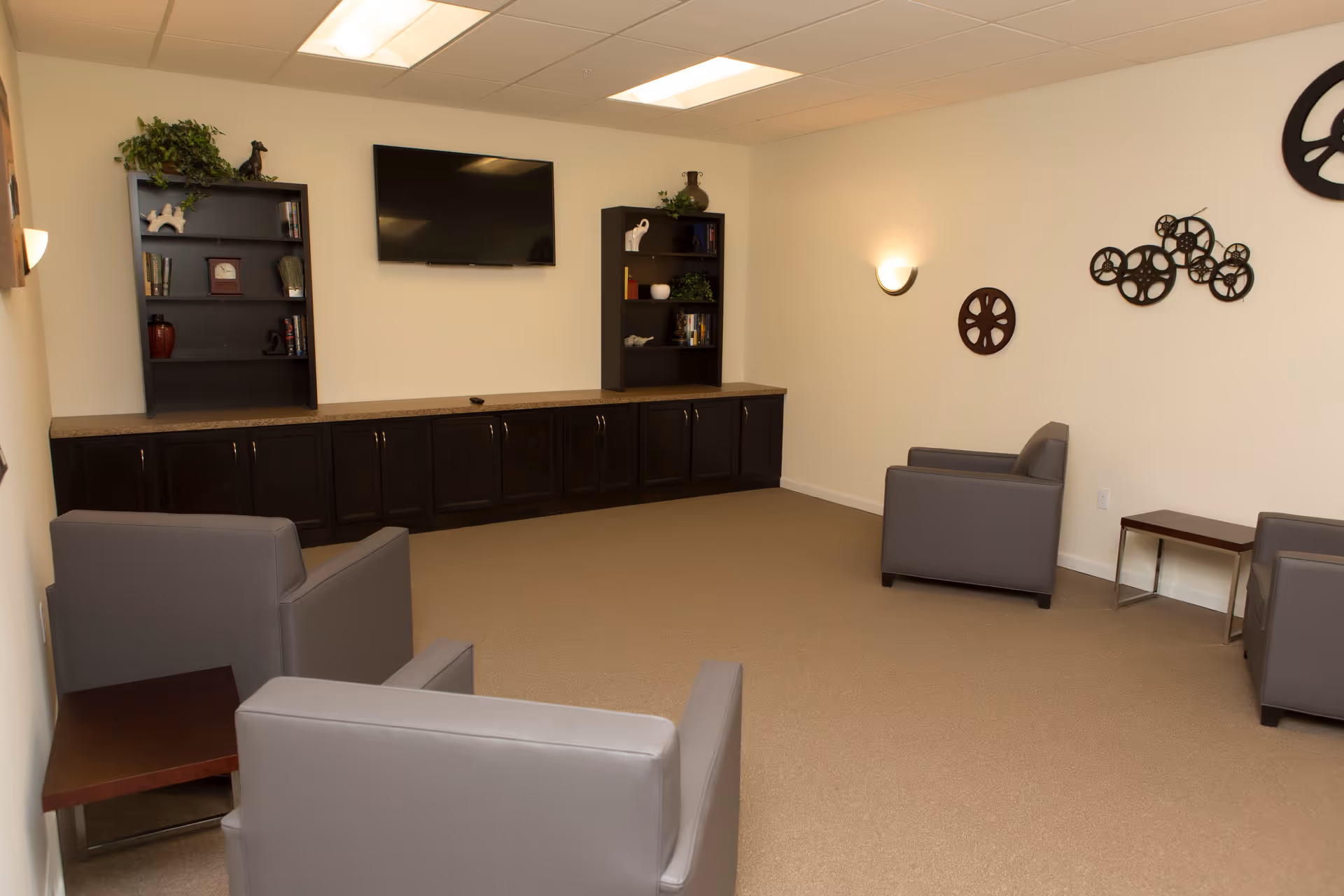 A cozy living room with beige walls and carpet, featuring four gray armchairs arranged around the perimeter. There are two dark wood bookshelves with decorative items and books, a flat-screen TV mounted on the wall, and wall-mounted lights. Decorative film reel wall art is displayed on one wall.