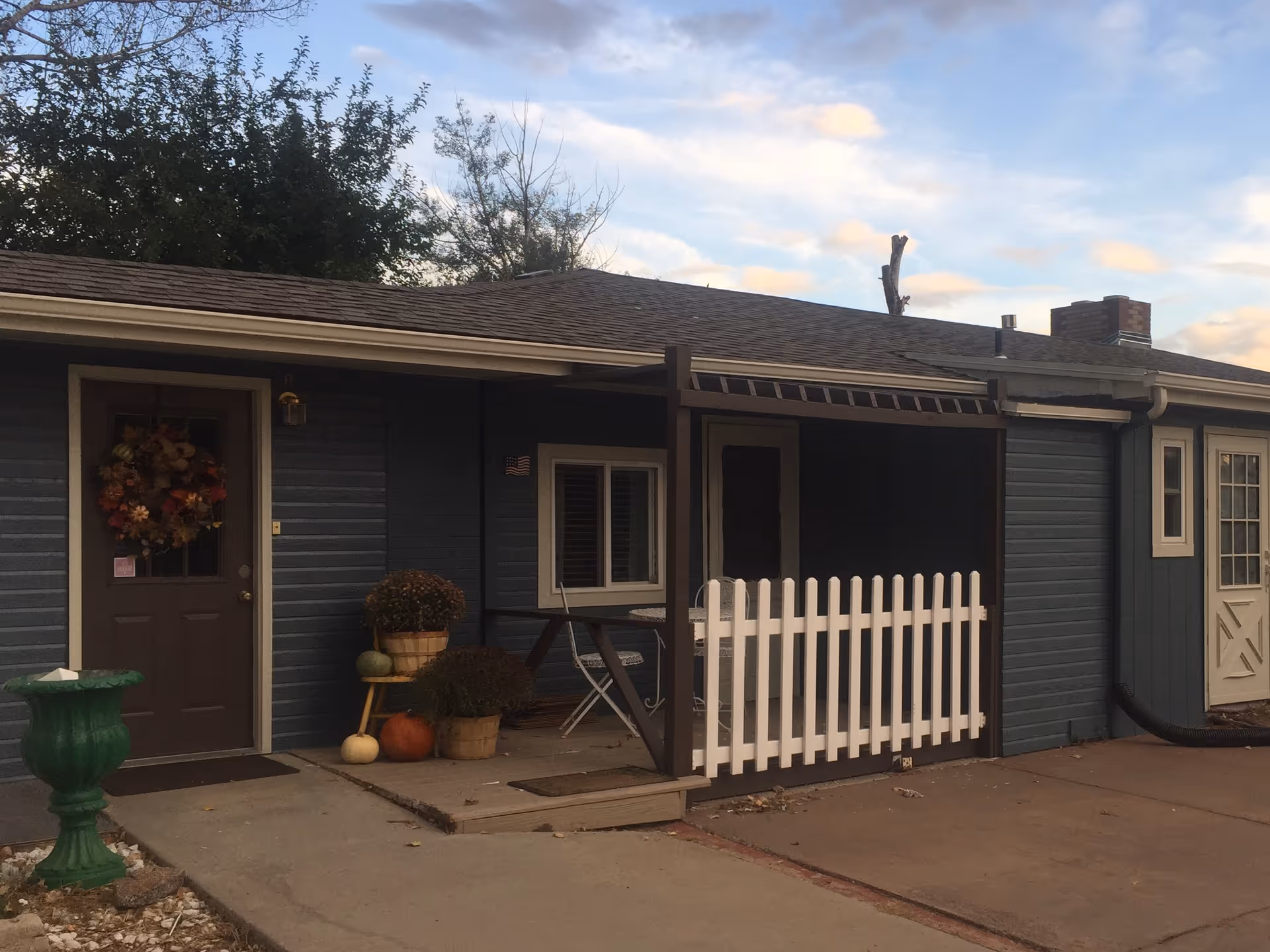 Exterior view of a single-story building with blue siding and a brown door decorated with a fall-themed wreath. There is a small porch area with a white picket fence, a table with two chairs, and several potted plants including pumpkins and flowers. The sky is partly cloudy with some trees visible in the background.