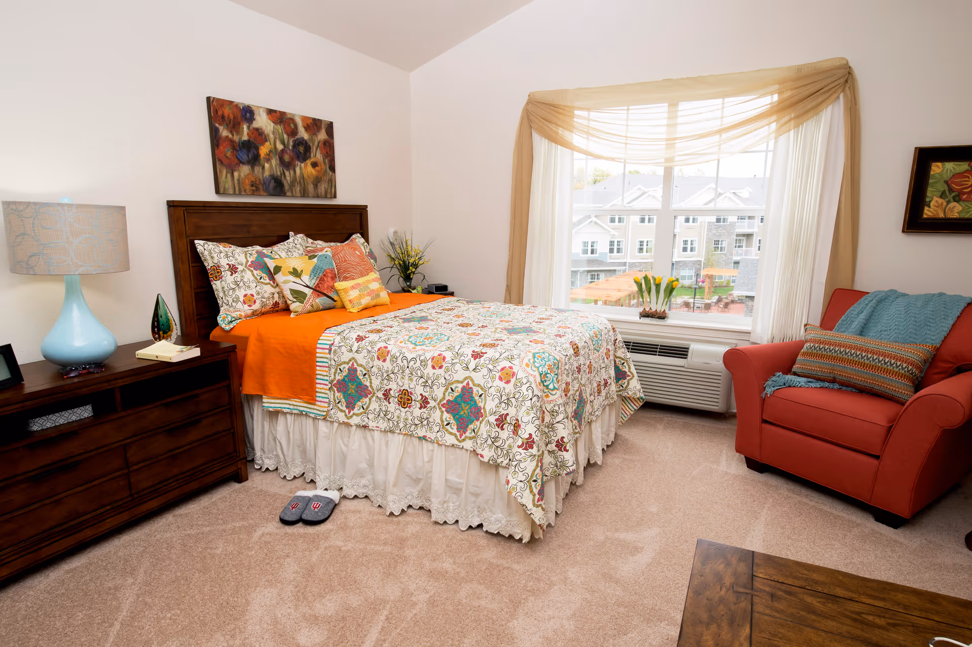 A cozy bedroom with a bed featuring a colorful floral and geometric patterned quilt and multiple decorative pillows. Next to the bed is a wooden nightstand with a blue lamp, a small decorative item, and a book. A pair of slippers is placed on the carpeted floor near the bed. There is a large window with sheer white curtains and a beige valance, letting in natural light and showing a view of an outdoor area with buildings. A red armchair with a blue throw blanket and a striped pillow is positioned near the window. The walls are white with a floral painting above the bed and another framed artwork on the wall near the armchair.