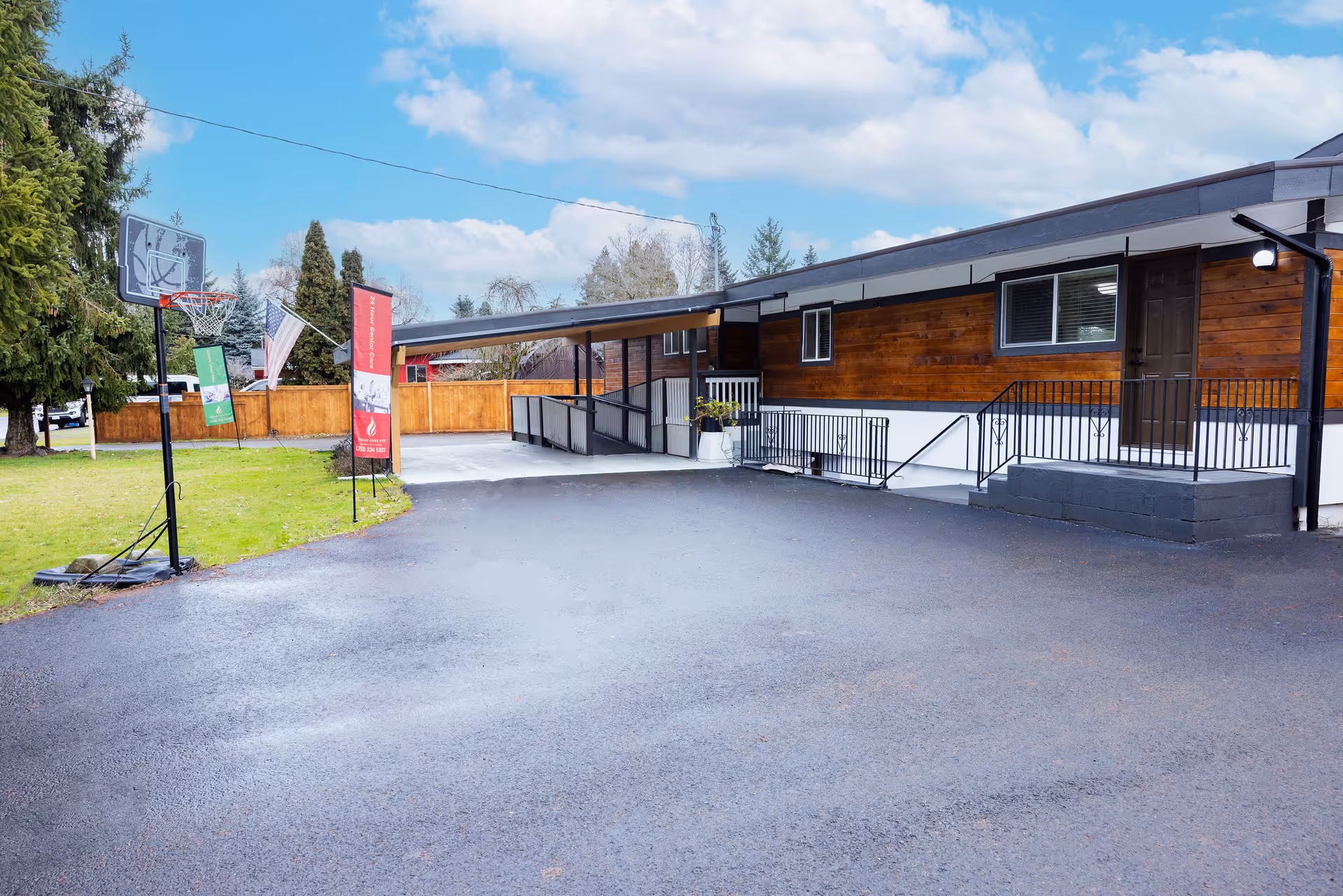 Front exterior of a single-story adult family home with a paved driveway, covered entry ramp, and a basketball hoop on the lawn.