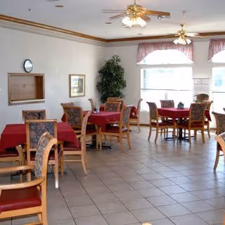 Bright dining room with several small tables covered in red tablecloths, wooden chairs, tile floor, ceiling fans, and large windows with valances.