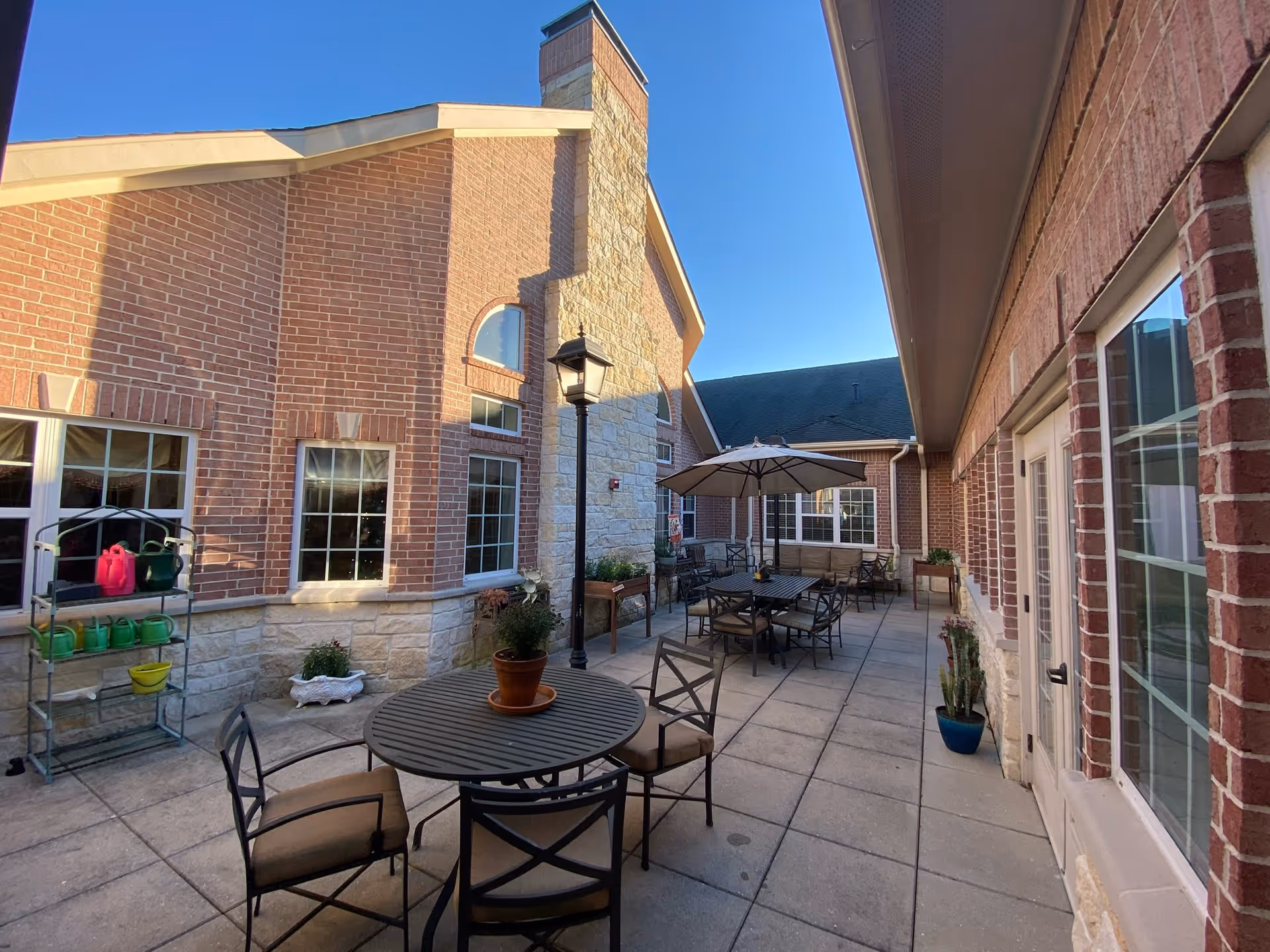 Outdoor patio area at The Heritage Memory Care facility with round tables and chairs, potted plants, a large umbrella, and brick and stone building walls under a clear blue sky.