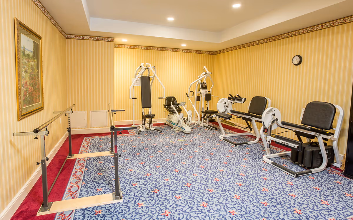 A senior living facility exercise room with yellow striped wallpaper and a blue patterned carpet. The room contains various exercise equipment including parallel bars, a seated chest press machine, a recumbent bike, and leg press machines. A framed painting hangs on the left wall and a clock is mounted on the right wall.