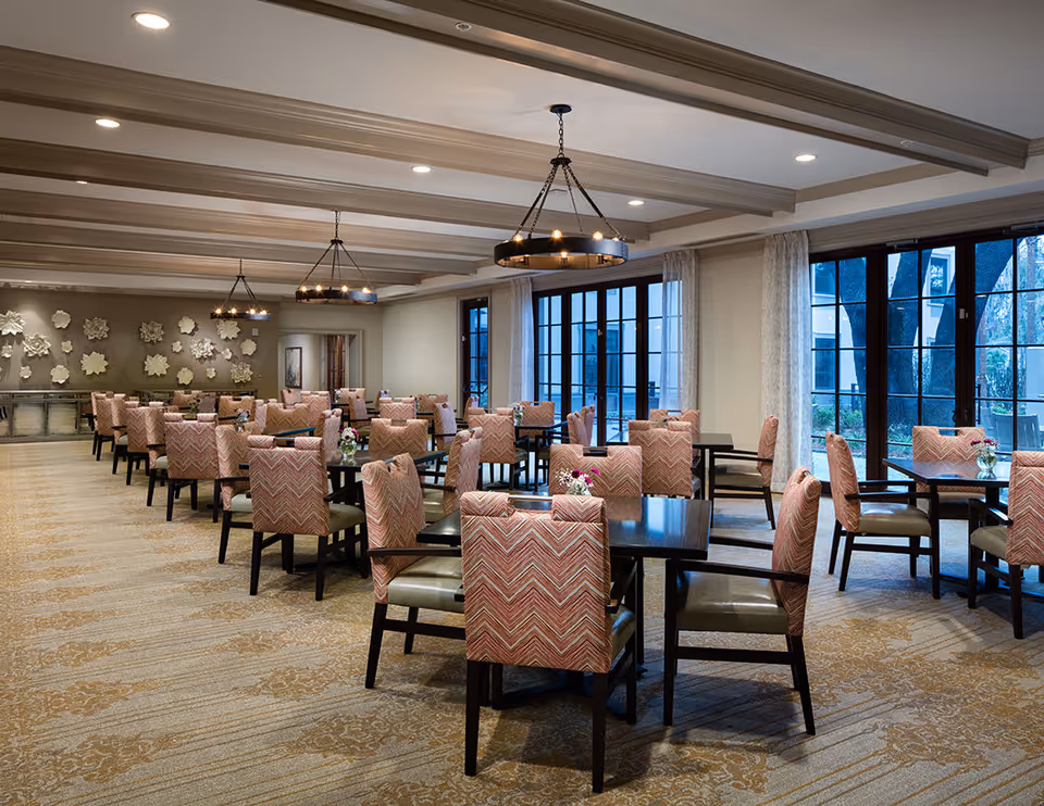 Spacious dining room with multiple dark wood tables, patterned upholstered chairs, large windows, and decorative chandeliers.