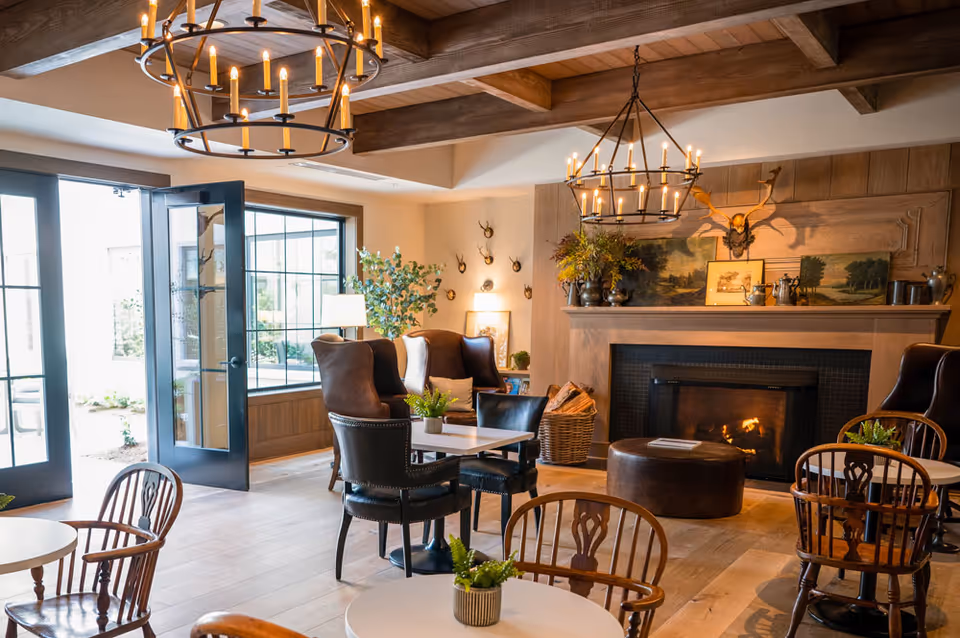 Cozy communal lounge with tables and chairs, leather armchairs, a lit fireplace, chandeliers, and open doors to the outside.