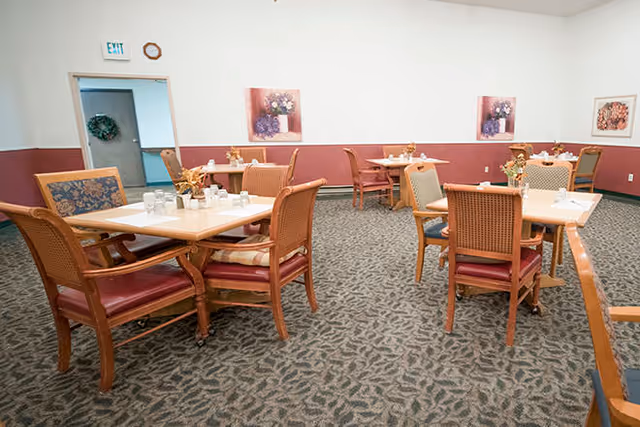 Empty dining room with wooden tables and chairs set on patterned carpet and floral artwork on the walls.