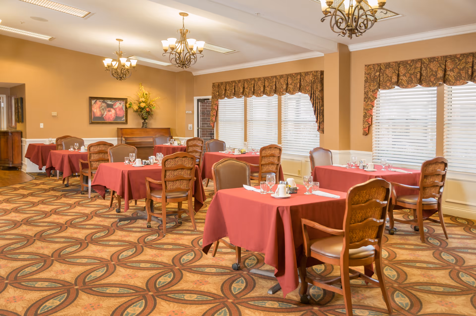A dining room with several tables covered in red tablecloths, each set with glasses, cups, and napkins. The room has patterned carpet flooring, beige walls, large windows with blinds and floral valances, and decorative chandeliers hanging from the ceiling. There is a piano and a floral arrangement against one wall.