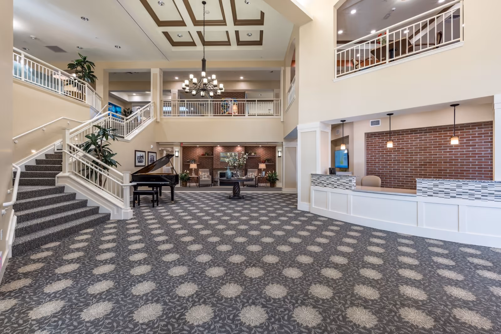 Spacious senior living facility lobby with patterned carpet, a grand piano, a seating area with chairs and a fireplace, a reception desk with pendant lights, and a staircase leading to an upper balcony.