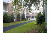 View of an outdoor pathway leading to the entrance of a brick and white building surrounded by green bushes and trees under a partly cloudy sky.
